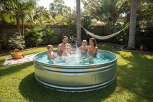 Load image into Gallery viewer, a family enjoying a stock tank pool in a sunny, lush backyard. The water is blue and crystal clea